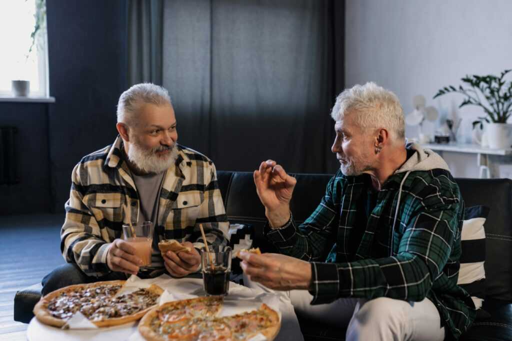 Two senior men enjoying pizza and drinks while chatting in a cozy indoor setting.